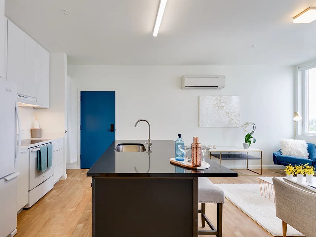 Modern kitchen and living area in Watercooler Apartments with a black island, white cabinets, and wood floors. A blue sofa, wall art, yellow flowers, blue front door, and large window complete this stylish space.
