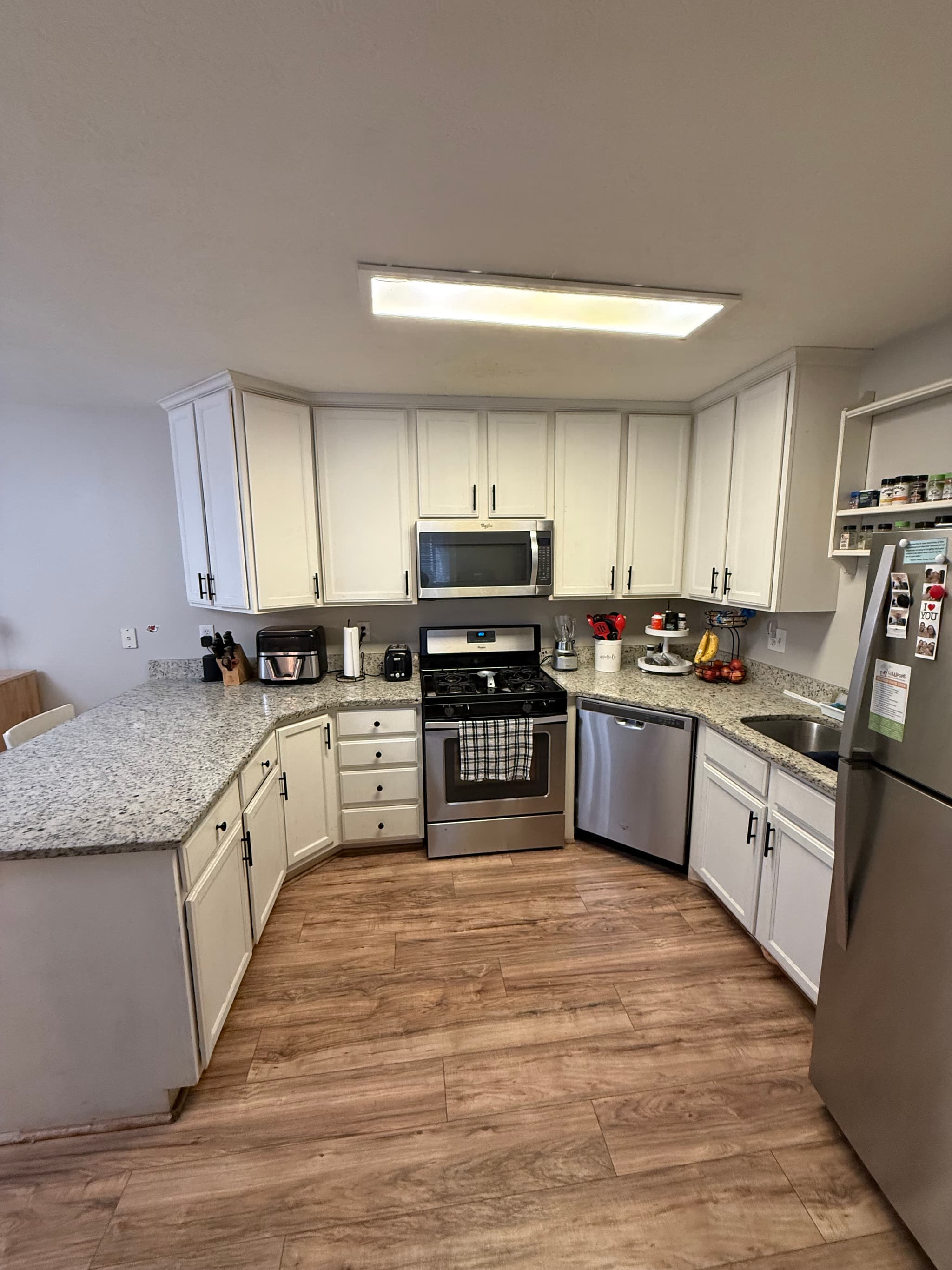 A modern kitchen at 282 W 960 S, APT C features white cabinets, granite countertops, stainless steel appliances, and wooden floors. Small kitchen appliances and utensils sit on the counters, with a towel hanging from the oven door.