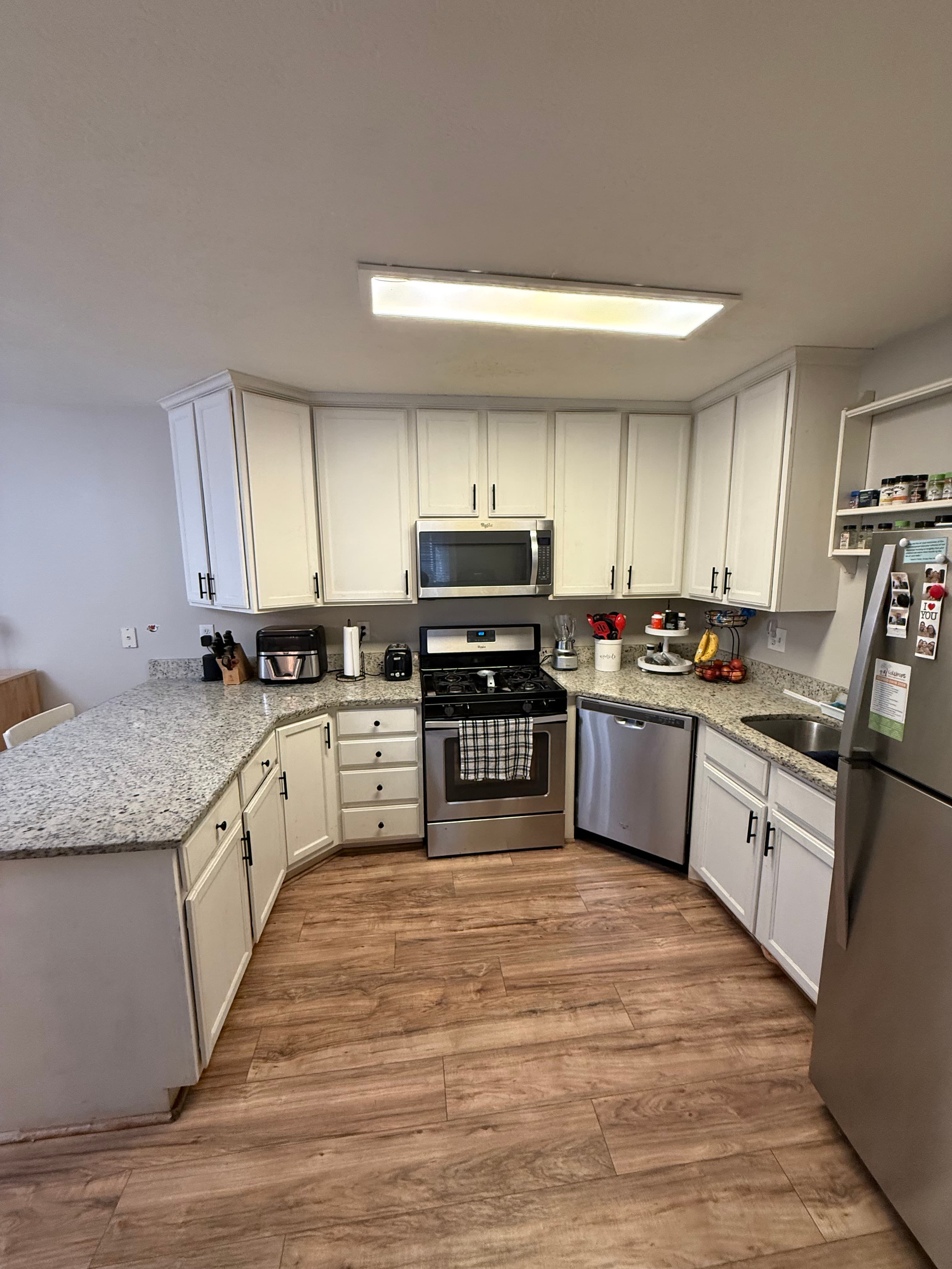 A modern kitchen at 282 W 960 S, APT C features white cabinets, granite countertops, stainless steel appliances, and wooden floors. Small kitchen appliances and utensils sit on the counters, with a towel hanging from the oven door.