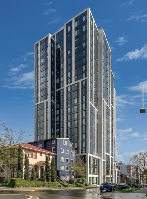 A modern high-rise building with glass and white panels, an architectural accolade, stands next to smaller, colorful houses on a sunny day with a blue sky. Cars are parked along the street, and trees line the sidewalk.