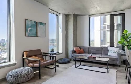 Modern living room with large windows, a gray sectional sofa, a brown chair, and elegant wall art—a space worthy of accolade. Bright, airy, and decorated with simple contemporary furnishings, it offers city and water views.
