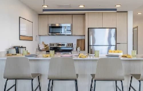 Modern kitchen with light wood cabinets, stainless steel appliances, and a white countertop. Four bar stools are arranged at the counter, with cups and yellow napkins—an accolade to stylish, functional design—placed in front of each seat.