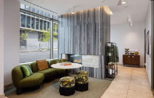 Modern Ace Berkeley lobby with green curved sofa, round table, two patterned stools, and a central double-sided fireplace. Large windows reveal a city street outside. Contemporary decor and neutral tones create a welcoming atmosphere.