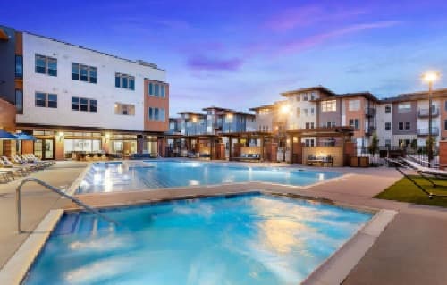 Modern Alas Over Lowry Denver apartment complex with a large outdoor swimming pool and a smaller hot tub in the foreground, surrounded by lounge chairs and lit by evening lights under a colorful twilight sky.