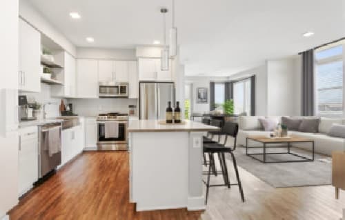Modern open-concept kitchen and living room at Alas Over Lowry Denver, featuring white cabinets, stainless steel appliances, a kitchen island with barstools, hardwood floors, and a bright, spacious seating area with large windows.