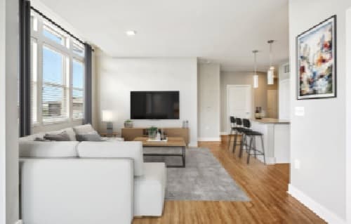 Modern living room with a light gray sectional sofa, wall-mounted TV, coffee table, and a gray rug at Alas Over Lowry Denver. An open kitchen with bar stools and pendant lights sits in the background while a large window lets in natural light.