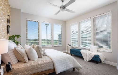 Bright bedroom with large windows, a neatly made bed with light bedding, and a blue bench with a white blanket. Neutral walls and a ceiling fan complete this modern, cozy space at Alas Over Lowry Denver, filled with sunlight.