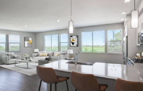 Modern open-concept living space at Alas Over Lowry Denver, featuring a kitchen island, two brown chairs, stainless steel appliances, and a bright living area with gray sofas and large windows overlooking greenery.