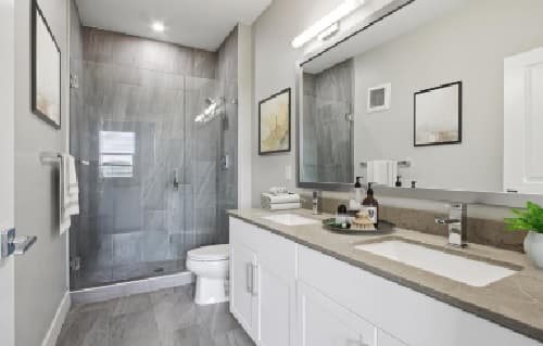 Modern bathroom at Alas Over Lowry Denver with a glass-enclosed shower, white cabinets, gray countertop with sink, wall-mounted mirror, toilet, towel racks, and framed artwork on light gray walls.
