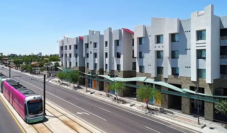 A street scene in Tempe shows a modern white apartment building with geometric features—possibly student apartments like Alight Tempe—beside a road where a pink and white light rail train travels. Trees line the sidewalk under a wavy green canopy.