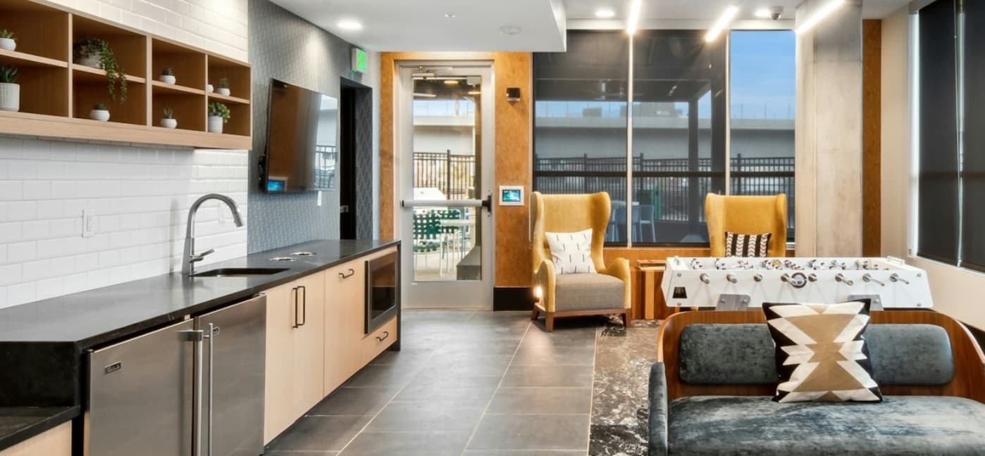 Modern lounge area at Amli Art District Denver with a kitchenette, open shelves, wall-mounted TV, two yellow armchairs by large windows, a foosball table, and a sofa with a geometric pillow. Bright and inviting with natural light.