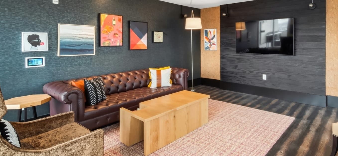 A modern living room at Amli Art District Denver with a brown leather sofa, armchair, wooden coffee table, wall-mounted TV, floor lamp, and colorful framed art on dark walls, featuring a pink patterned rug and wood flooring.