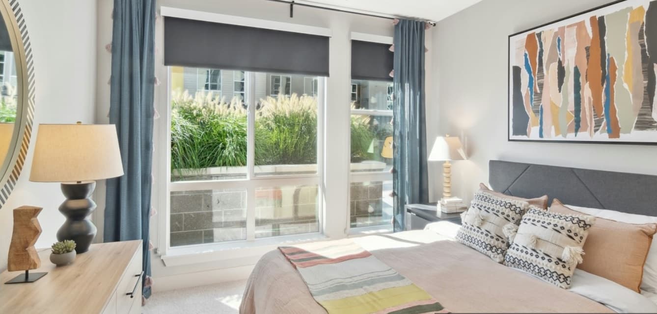 Bright bedroom at Amli Art District Denver with large windows, a neatly made bed with patterned pillows, light wood furniture, abstract wall art, two lamps, and blue curtains. Sunlight streams in, illuminating the cozy, modern decor.