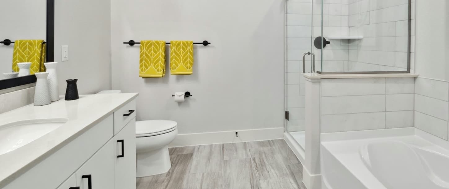 Modern bathroom at Amli Art District Denver with white cabinets, a sink, toilet, glass-enclosed shower, and bathtub. Two yellow patterned towels hang on a black towel bar. Light gray floor tiles and white walls create a clean look.