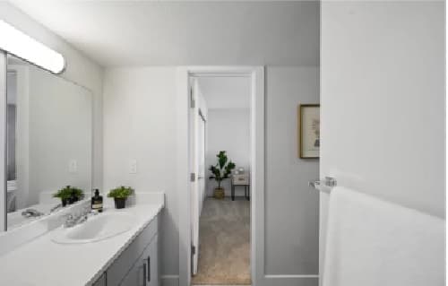 A modern, white bathroom at The Alpine Apartments Fort Collins features a single sink, large mirror, green plants on the counter, and a white towel on a rack. Through the open door, an office with a plant and desk is visible.
