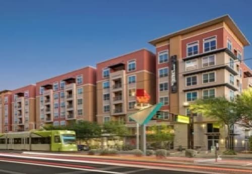 A modern multi-story apartment building, The District on Apache Tempe, features balconies and large windows beside a street with blurred traffic and a passing green light rail train in the evening. Trees and streetlights line the sidewalk.