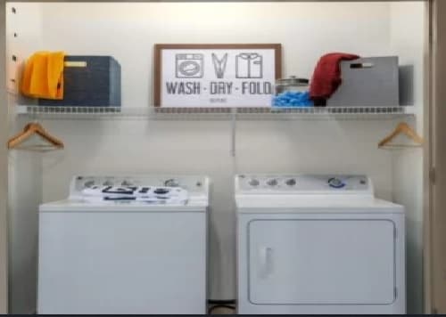 A laundry room at The District on Apache Tempe features a washing machine and dryer, two storage bins, folded towels, hangers on a shelf, and a framed sign that says Wash - Dry - Fold above the appliances.