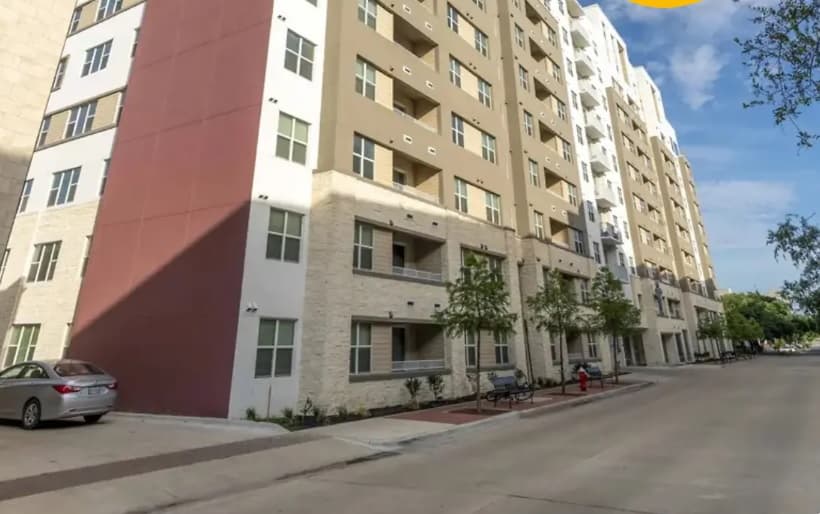 A modern multi-story apartment building, perfect for student housing in College Station, with balconies, beige and white exterior walls, tree-lined sidewalks, and a parked silver car along a quiet street on a sunny day—welcome to Yugo Apex.