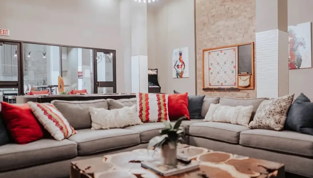 A modern living room at Yugo Apex features a large gray sectional sofa with red, white, and patterned pillows, a wood coffee table, wall art, and a game board mounted on a brick accent wall—perfect for College Station living.