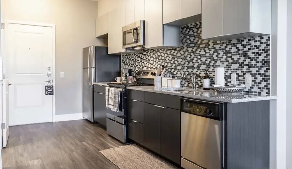 Modern kitchen at Yugo Apex in College Station with stainless steel appliances, dark lower cabinets, light uppers, and a black-and-white mosaic tile backsplash. A countertop holds essentials while a rug decorates the wood floor.