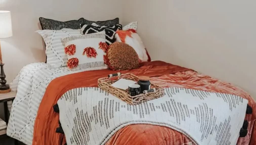A cozy bed at Yugo Apex, College Station with white and orange bedding, decorative pillows, and a tray holding mugs and candles. The bed is beside a small lamp against a neutral wall, creating a warm, inviting atmosphere.