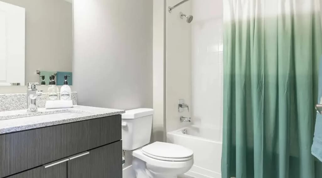 Modern bathroom in Yugo Apex student housing, College Station, with a white toilet, bathtub and green ombre shower curtain, granite countertop sink, mirror above the sink, light-colored walls, and a towel hanging on the right.