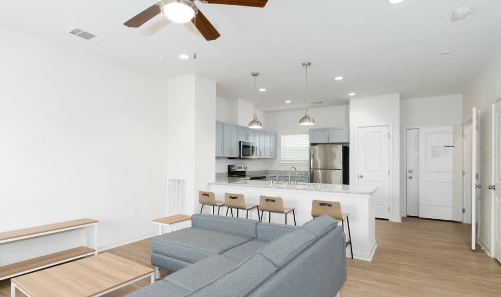 Modern open-concept living space in Aspen Heights student housing, featuring a gray sectional sofa, wooden coffee table, stylish kitchen with light blue cabinets, stainless steel appliances, breakfast bar with four stools, ceiling fan, and wood flooring.