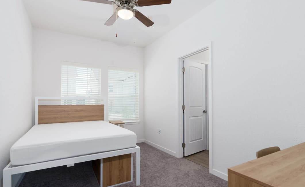 A simple, clean bedroom in Aspen Heights student housing with a bed frame and mattress, a wooden desk and chair, carpeted floor, ceiling fan, two windows with blinds, and an open door leading to another room.
