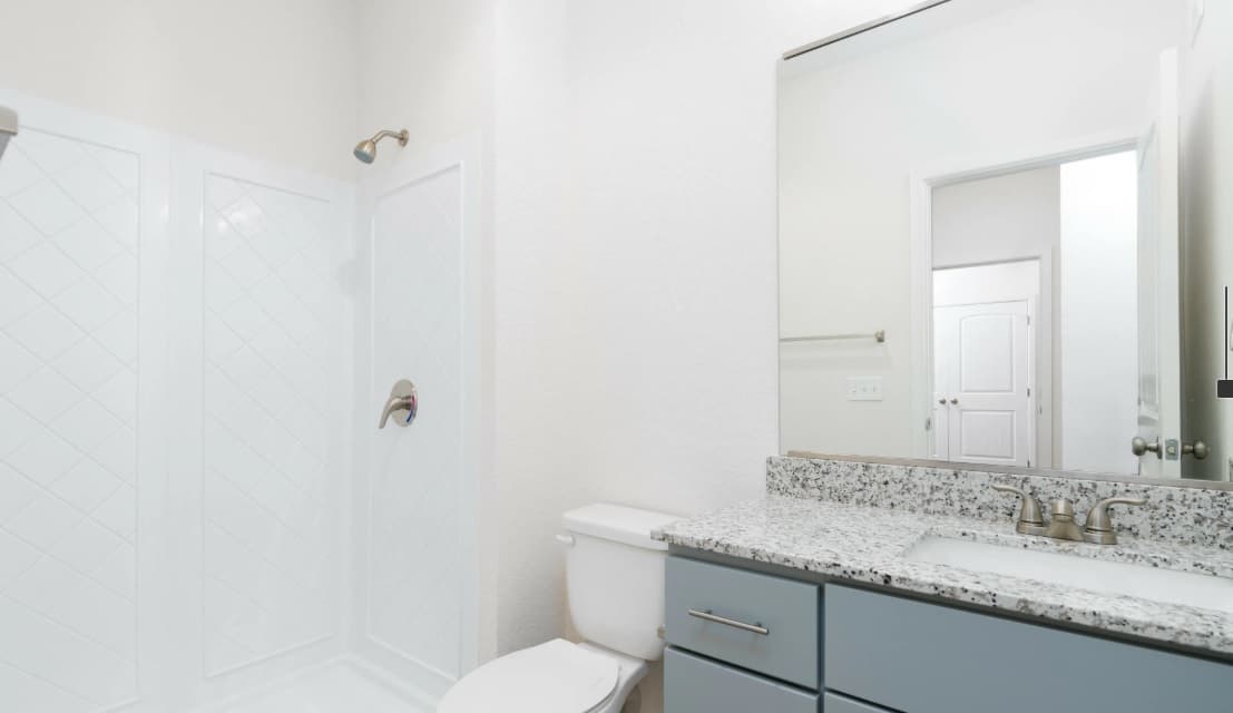 A clean, modern bathroom in Aspen Heights student housing features a glass shower, white toilet, and granite countertop with a sink and large mirror above blue cabinets. The well-lit space has neutral walls, perfect for stylish apartments.