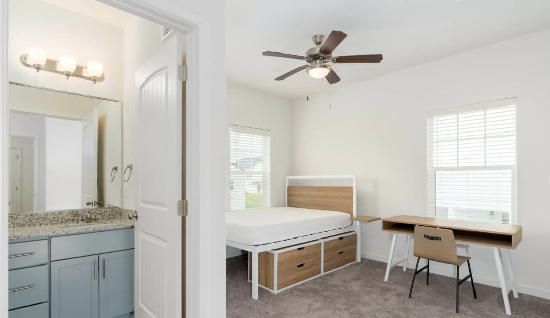 A bright Aspen Heights bedroom with a ceiling fan, bed with storage drawers, wooden desk and chair, beige carpet, plus an adjoining bathroom featuring a granite countertop and light blue cabinets.