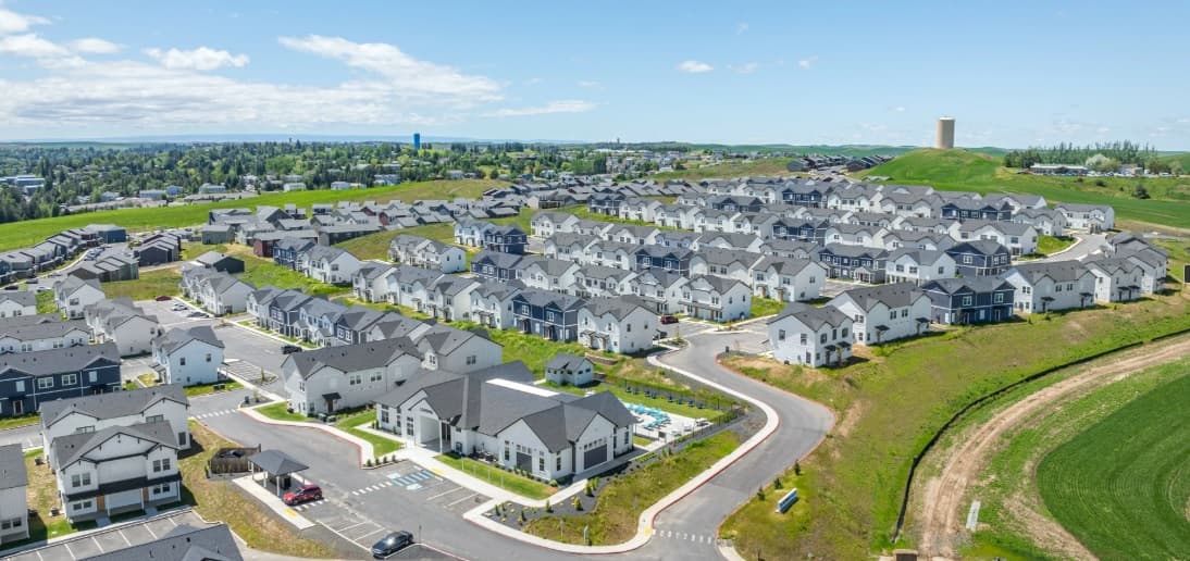 Aerial view of a suburban residential neighborhood with many modern, white and gray Aspen Heights apartments, curved streets, green lawns, and open grassy areas under a blue sky with scattered clouds.
