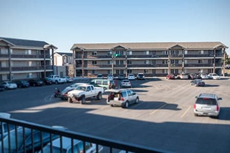 A large, paved parking lot with several parked cars sits in front of Campus Park Rexburg, a three-story apartment complex with balconies and tan exteriors on a clear, sunny day.