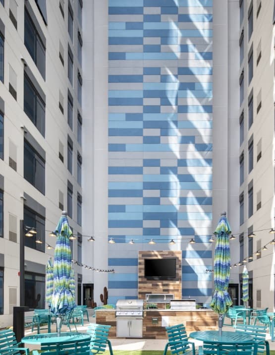 A modern outdoor courtyard at Atmosphere Tempe features teal chairs, blue-green striped umbrellas, a grill, and a TV. The space sits beside a tall building wall with blue rectangular patterns, flanked by two white buildings.