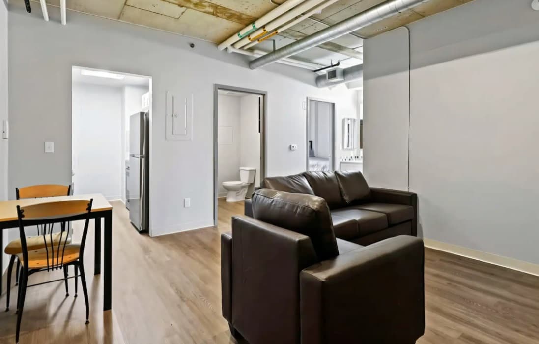 Modern apartment interior at Auraria Lofts featuring a brown leather sofa and armchair, small dining table with chairs, visible kitchen appliances, bathroom, exposed pipes, and a light wood floor.