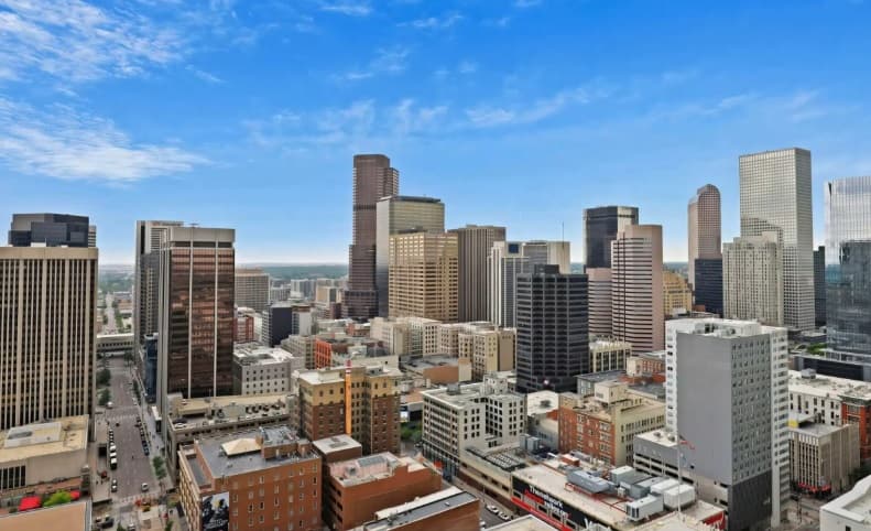 Aerial view of Denver's city skyline featuring tall modern skyscrapers, nearby apartments like Auraria Lofts, and smaller buildings under a bright blue sky with scattered clouds.