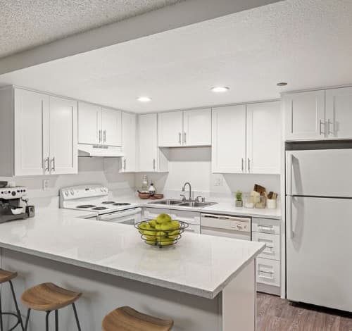 Modern kitchen at Aztec Campus Apartments with white cabinets and appliances, a white countertop with a bowl of green apples, three wooden bar stools, and a coffee machine; the space is brightly lit with recessed lighting.