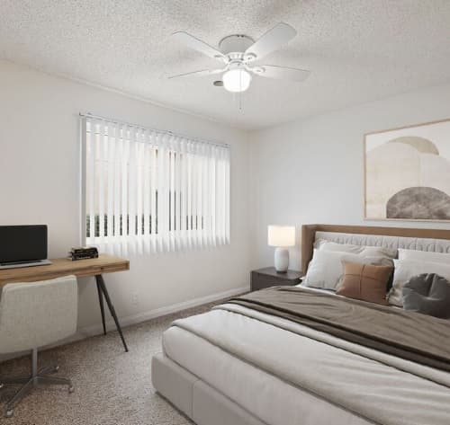 A modern, minimalist bedroom at Aztec Campus Apartments features a large bed, neutral bedding, a wooden desk with a computer, a chair, nightstand with lamp, abstract wall art, vertical blinds on the window, and a ceiling fan with light.