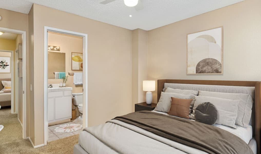 A cozy Aztec Campus Apartments bedroom features a neatly made bed, beige walls, a nightstand with a lamp, and an abstract painting. An open door reveals a bathroom with a white vanity and mirror, plus another room down the hall.