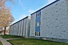A three-story apartment building with a stone-patterned façade, blue panels, and many windows stands beside a sidewalk and grassy lawn on Campus Way Provo under a clear sky.