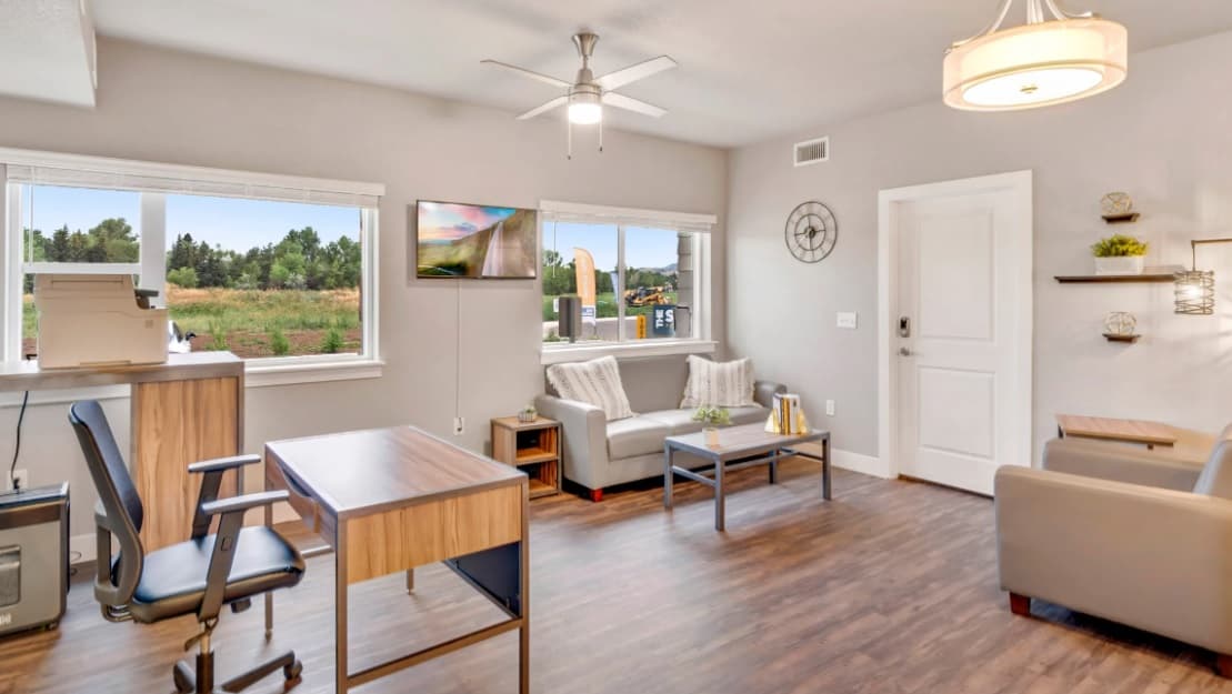 A modern office at Bighorn Landing Fort Collins features wood flooring, two desks and chairs, a printer by the window, gray sofas, a coffee table, wall-mounted TV, large windows with outdoor views, and neutral-colored decor.