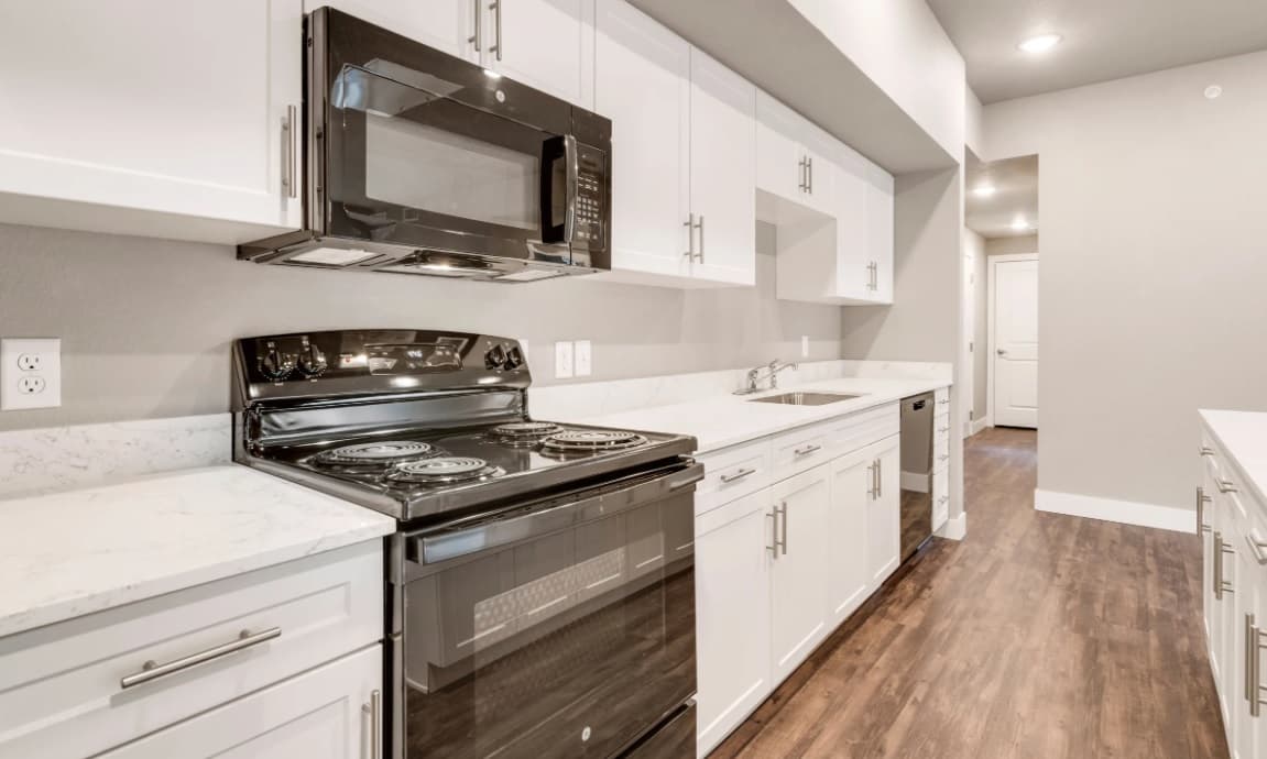 Modern kitchen at Bighorn Landing Fort Collins with white cabinets, black electric stove and microwave, white marble countertops, stainless steel sink, dishwasher, wood-style flooring, and well-lit recessed ceiling lights.
