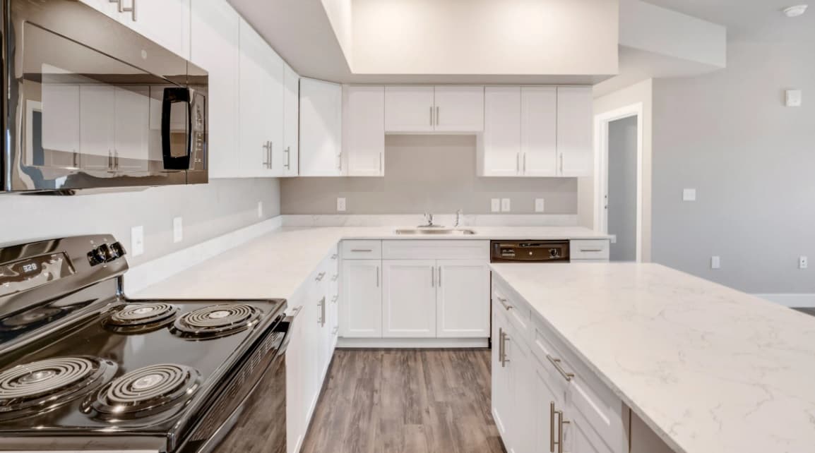 Modern kitchen at Bighorn Landing Fort Collins with white cabinets, marble countertops, stainless steel appliances, and wood-style flooring. Features an electric stove, microwave, dishwasher, and a sink under a cabinet with neutral-toned walls.
