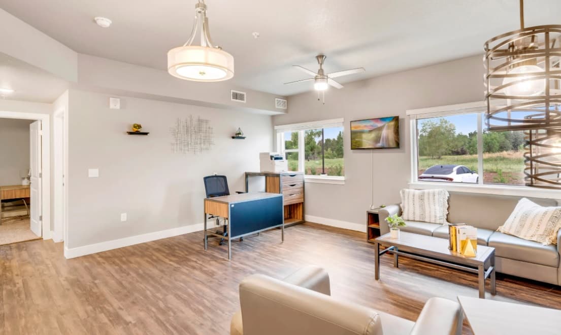 A bright office space at Bighorn Landing Fort Collins with a wooden floor, modern furniture, a desk with a chair, a coffee table, and two sofas near large windows showing greenery and a parked car outside. Stylish light fixtures hang from the ceiling.