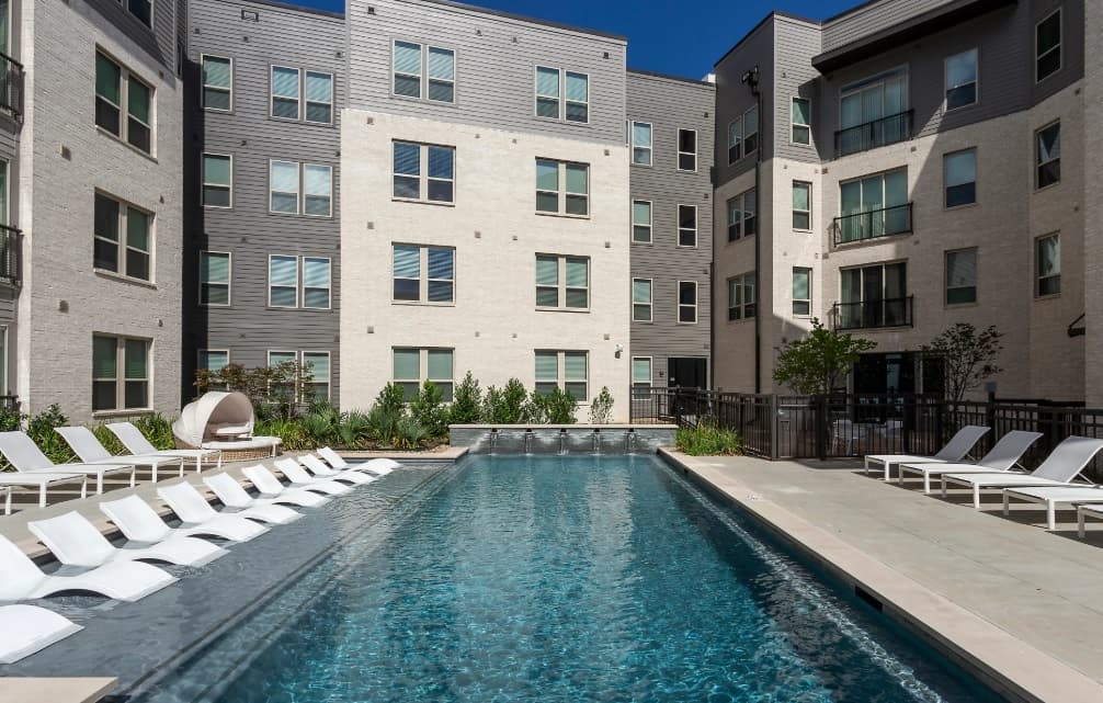 A modern apartment complex at Berkeley House in College Station surrounds a rectangular outdoor pool with lounge chairs along both sides and a cabana seat in the corner, all under a clear blue sky.
