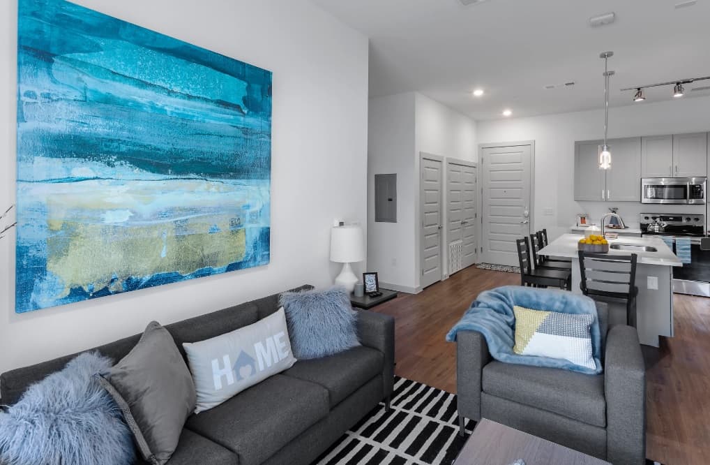 Modern living room at Berkeley House with gray sofas, blue and gray pillows, a large blue abstract painting, and a striped rug. Open kitchen features white cabinets and stainless steel appliances—perfect student housing in College Station.