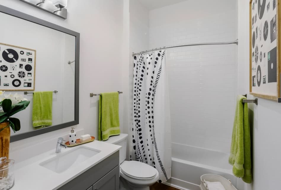 Modern bathroom at Berkeley House with white walls, a bathtub with a black and white shower curtain, gray vanity, large mirror, green towels on racks, and framed art. A plant and soap dispenser sit on the sink—a stylish College Station retreat.