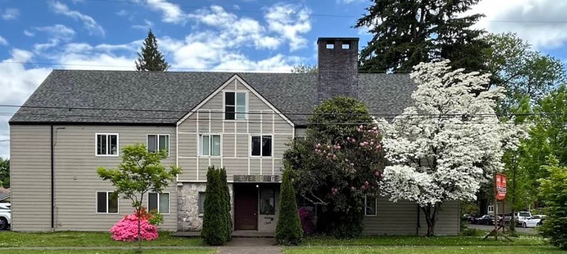 A two-story beige apartment building with a gray roof, central chimney, and stone entryway sits amid blooming trees and shrubs with white and pink flowers, creating a setting reminiscent of a peaceful wildlife habitat under a partly cloudy sky.