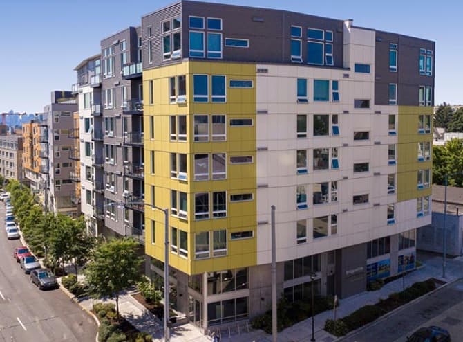 Modern mid-rise apartment building with yellow and gray exterior panels, large windows, and street-level retail space. Cars are parked along the tree-lined sidewalk on a clear day—ideal for those seeking Seattle apartments like Bridges@11th.