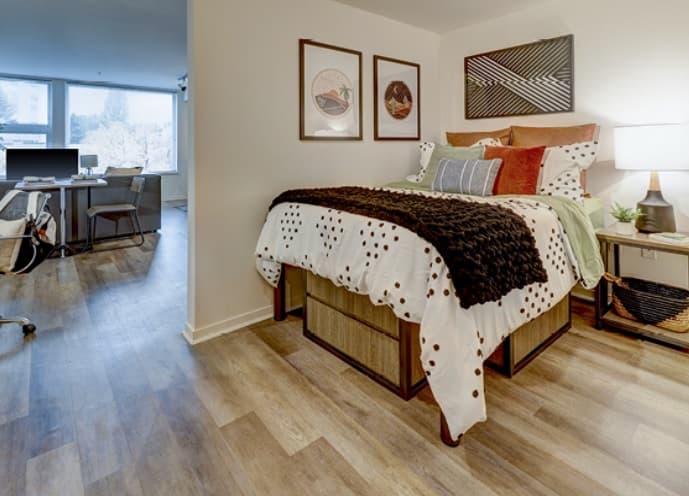 A cozy bedroom at Bridges@11th features a bed with polka dot bedding, decorative pillows, and a chunky blanket. Art hangs above the bed, while a lamp and plant rest on the nightstand. An adjacent room with a desk and windows is visible.