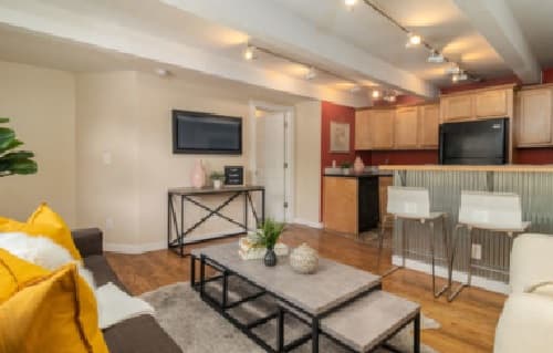 Modern living room and kitchen at Buffalo Canyon Apartments Boulder feature wood floors, beige and red walls, a brown sofa with yellow pillows, two bar stools at a counter, wall-mounted TV, and decorative plants on the coffee table and console.